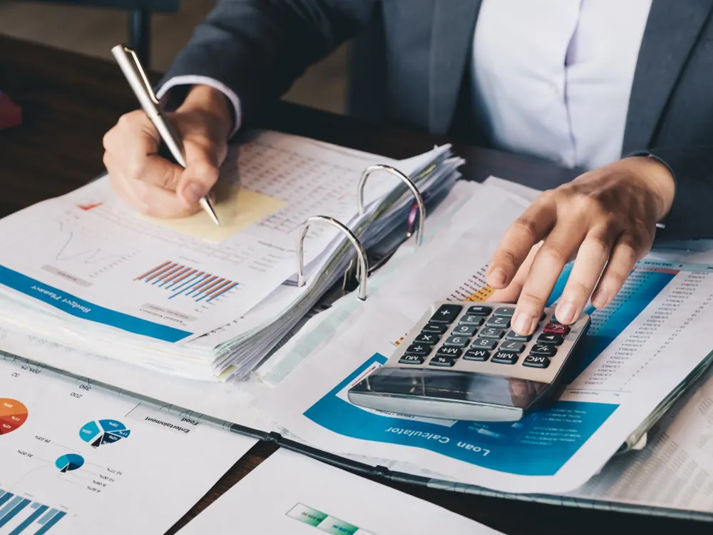Hand writing in a ledger beside a calculator and financial reports